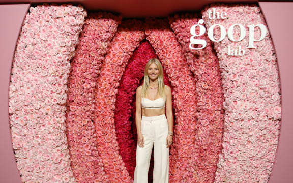A blonde woman with white pants and a white bra top stands in front of a pink flower wall.