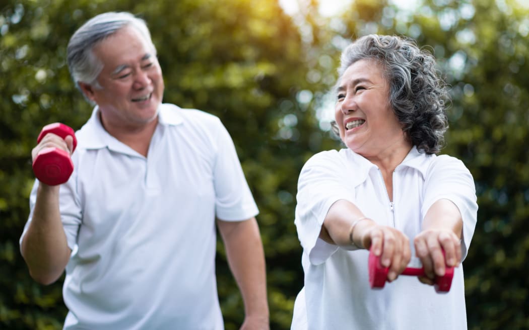 Asian Senior Couple exercising with red dumbbells at the outdoor park togetherness. Smiling Chinese or Thai or Japanese people.