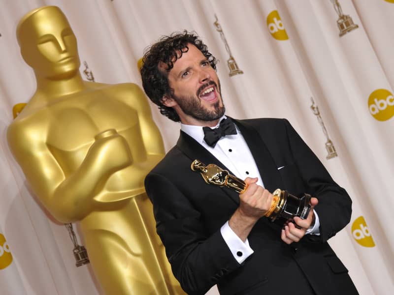 Bret McKenzie makes a zany face and holds an Academy Award, as he stands in front of a giant gold Oscar statuette.
