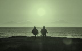 The image is of a pair of people in the foreground on a beach, watching the sun go down. Both are sitting on folding chairs.
