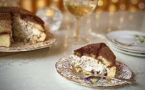A whole Zuccotto (a dome of sponge filled with whipped cream laced with chocolate and hazelnuts) sits on a plate with a wedge cut out of it, the wedge is on a plate in the right foreground. There is a glass of prosecco and fairy lights in the background.