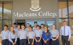 A group of students stand in front of a building with MACLEANS COLLEGE written on the front in silver lettering. The students all wear a navy and white striped shirt and navy pants or skirts. Their principal stands to the right wearing a shirt and tie. They all smile.
