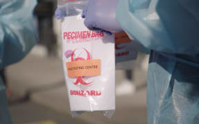 A close up of a person wearing rubber gloves and a blue plastic gown holding a ziploc bag with the words "Specimen bag from testing centre biohazard" printed on it.