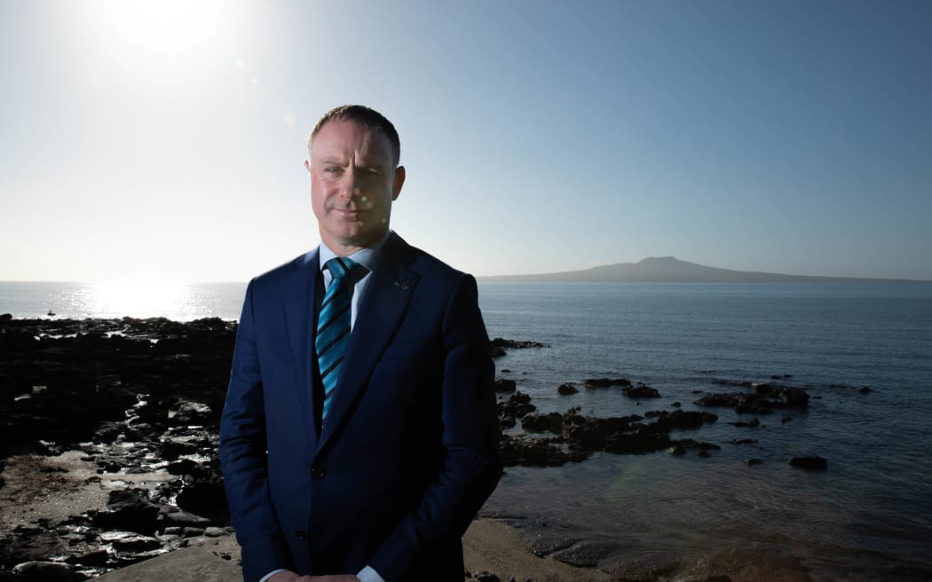 Climate Change minister Simon Watts, wearing a blue suit, stands in bright sunlight near a rocky shore, with Rangitoto Island in the background.