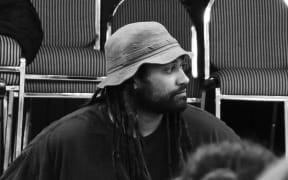 Te Kahu Rolleston is sitting on a school stage in front of a group of students. He is wearing a hat that is almost but not quite a bucket hat and looking to his left. The image is in black and white.