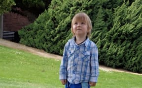 A young boy with shaggy brown hair is looking up at the sky. He is in a backyard, standing on grass, with a dense hedge behind him. He is wearing a blue checked shirt.