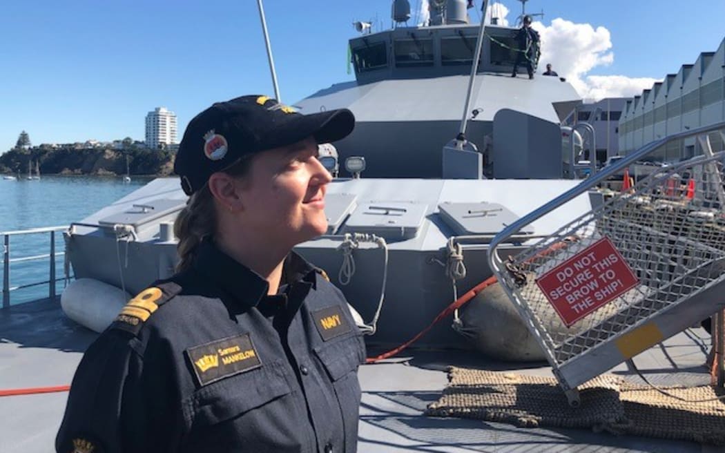 Samara smiles off to the right. Behind her is a naval vessel. It is a bright sunny day.