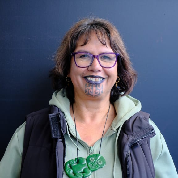 Te Pā o Rākaihautū Chairperson Rangimarie Parata-Tukurua stands smiling at the camera. She wears purple glasses and a hei tiki hangs around her neck.