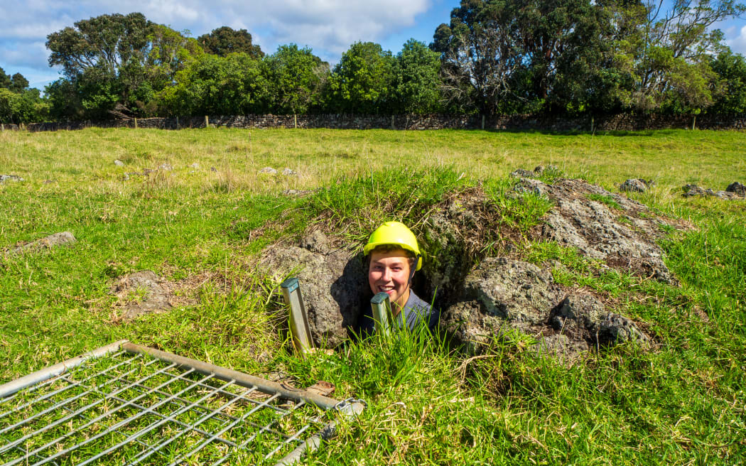 A man in a hard hat standing on a ladder in a hole in a grassy field. The sky is blue but cloudy and there is a metal grate on the ground in front of the hole. The man is smiling.