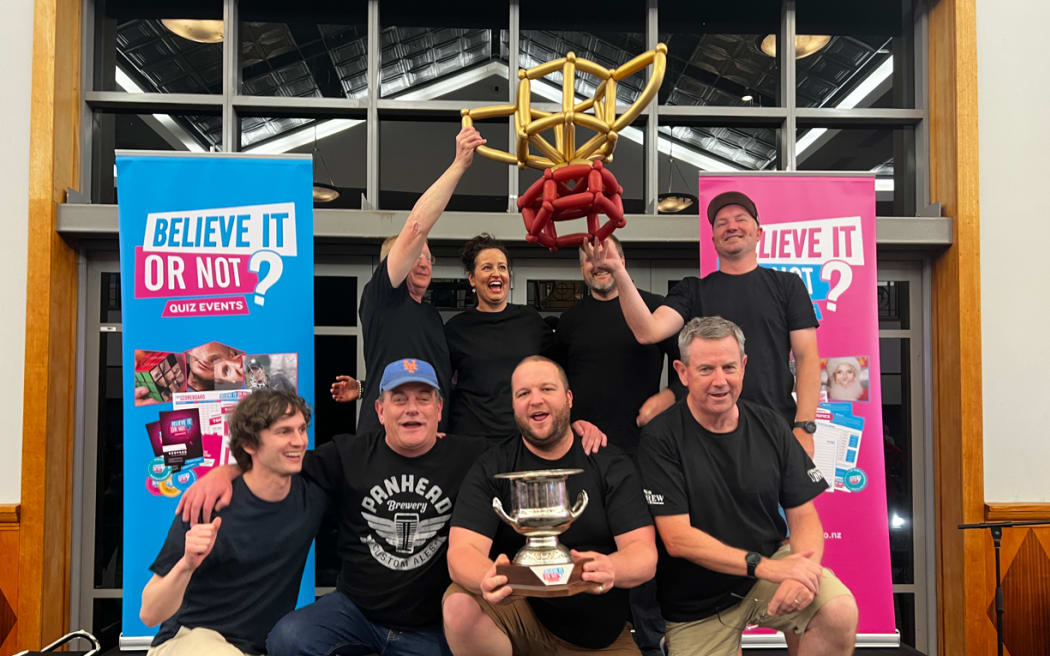 A pub quiz team dressed in black pose for a photo, some kneeling, some standing, holding aloft their trophy. They all smile.