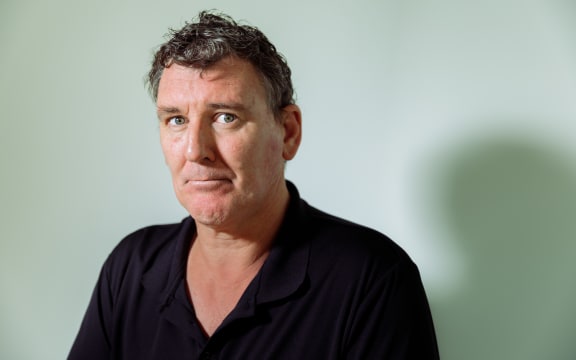 Jeremy Buxton sits for a photograph, his shadow being cast on the wall behind him.