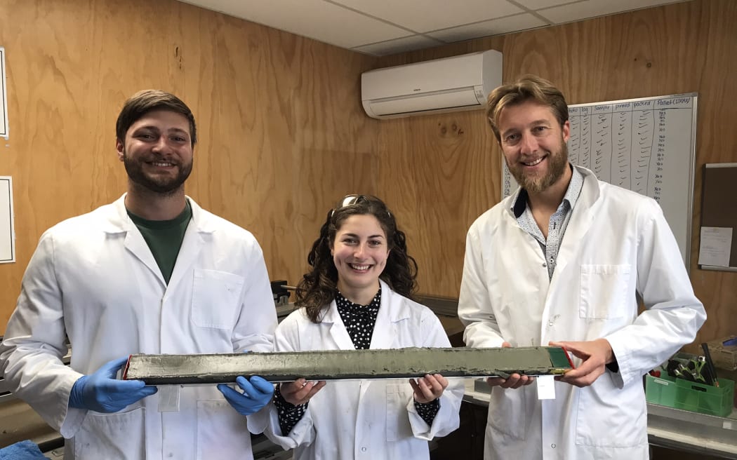Three people in labcoats are smiling and holding a 90 cm core segment with olive-green mud in it.