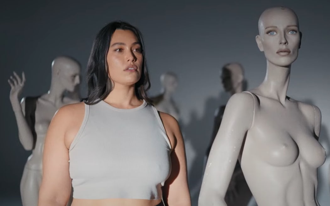 Isabella Moore - a woman with long brown hair and a grey tank top - stands among a group of thin mannequins.