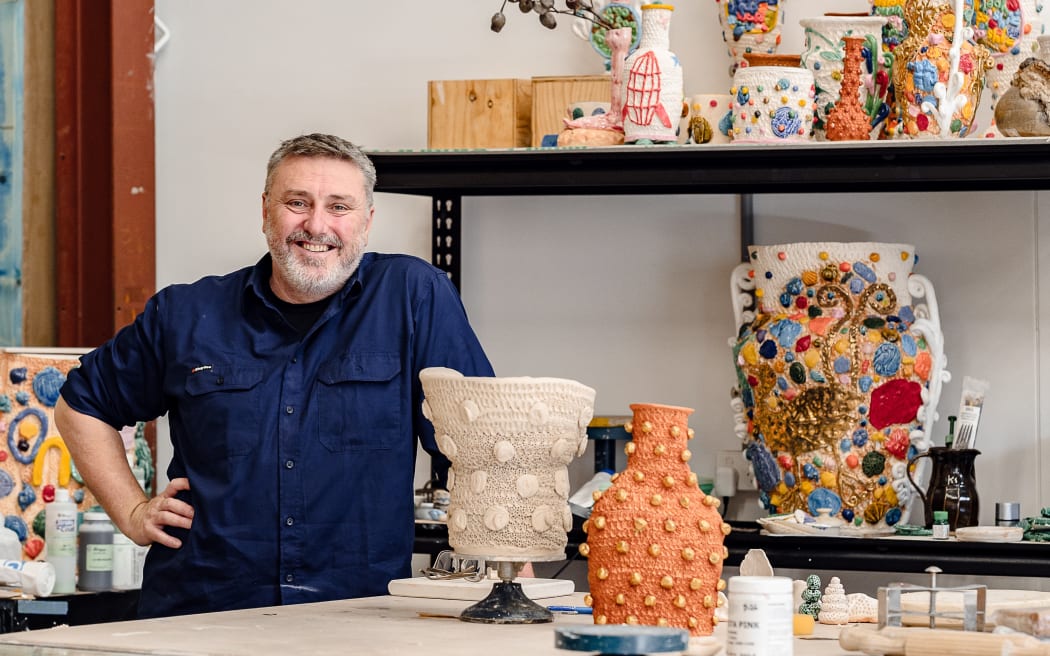 Glenn stands by a table covered in ceramics. He smiles.