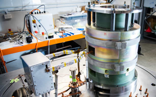 A stack of cylinders connected to a small metal box on a metal table. In the background is a metal lab bench with tools and equipment scattered around.