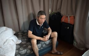 Gao Litong sits on his bed which is a mattress on the floor of a living room which he shares with another Chinese worker who came to New Zealand to work.