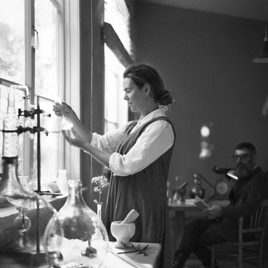 Kate stands in a dimly lit studio by a table of distilling equipment, holding a small glass vial to the light. David sits at a table in the background.