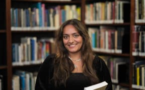 Anjuli smiles at the camera. She is standing in a library, holding a book. She wears a black top.