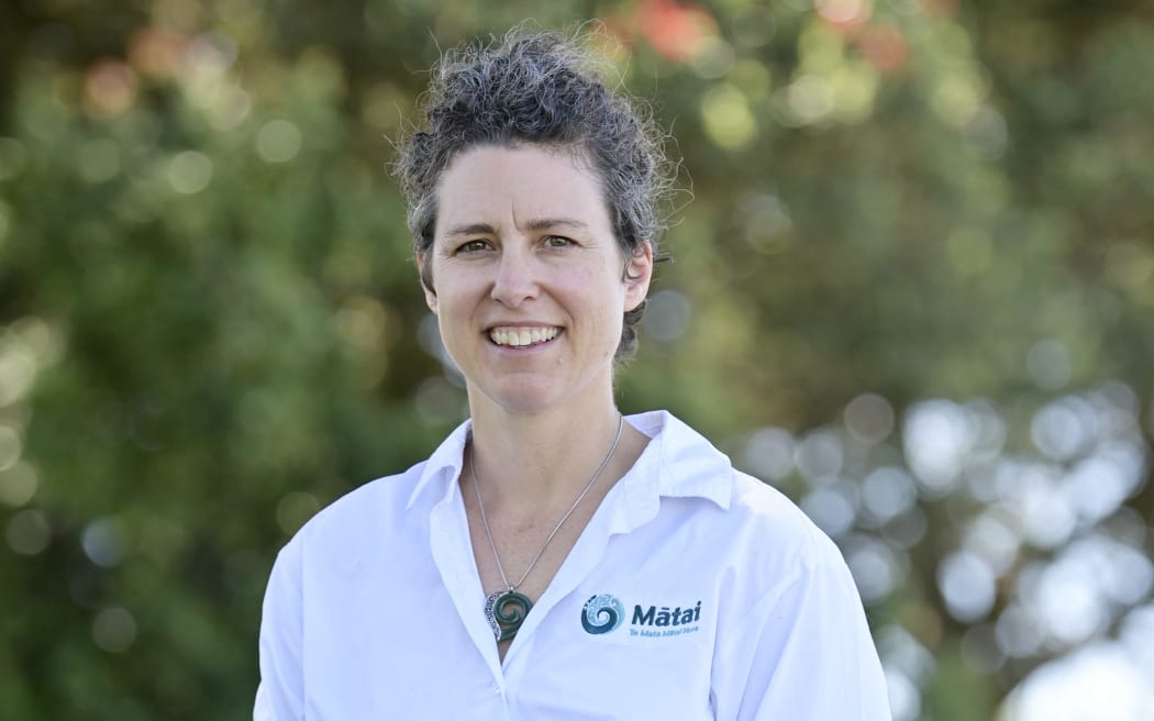 A woman wearing a white lab coat with a logo for Mātai Medical Research Institute on it stands outside, smiling at the camera.