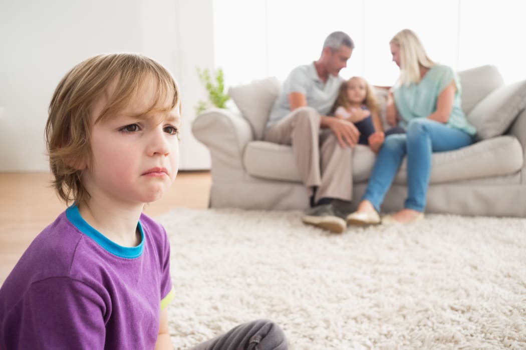 Photo of an upset boy sitting on floor while parents enjoying with sister on sofa at home