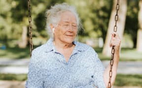 A senior woman sits on a swing smiling as she listens to music through wired headphones.