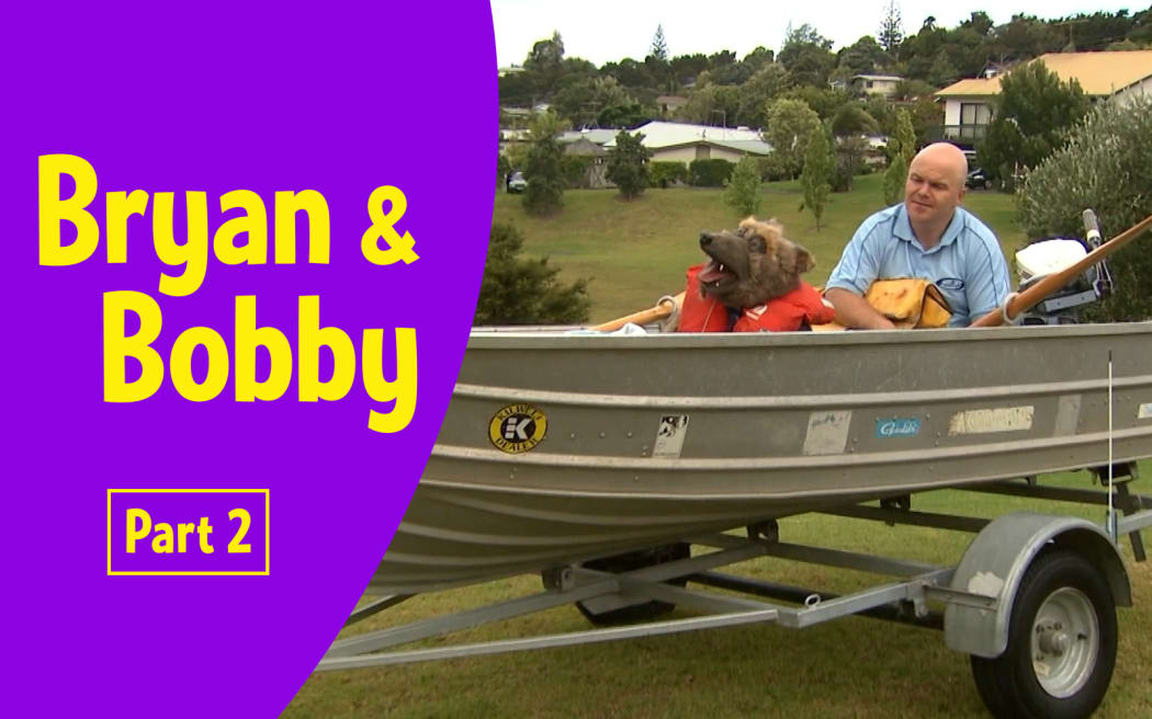 Photograph of Bryan (male adult) and Bobby (realistic large dog puppet) in a small boat which is still in its trailer on the grass
Text reads   "Bryan and Bobby Part 2”