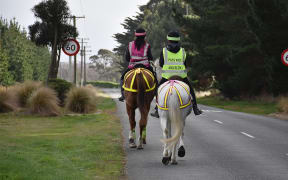 Two horseback riders riding down a paved country road wearing hi-vis vests wit 'PASS SLOW AND WIDE' written on the back.  There is a sixty kilometre per hour sign in the background.