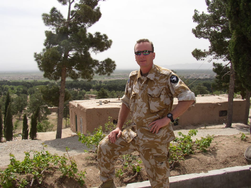 Army veteran Bill Blaikie overlooking the destroyed division base in Herat.  A man in army uniform stands with one leg on a clay wall.  In the background is are mud brick buildings and pine trees