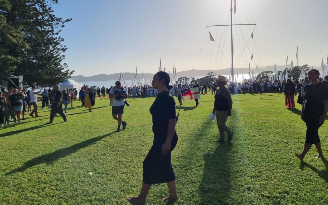 A dramatic soundscape is being played as the people holding white flags disperse, walking around the manuhiri side of the wharenui and atea, in this part pōwhiri, part art piece.