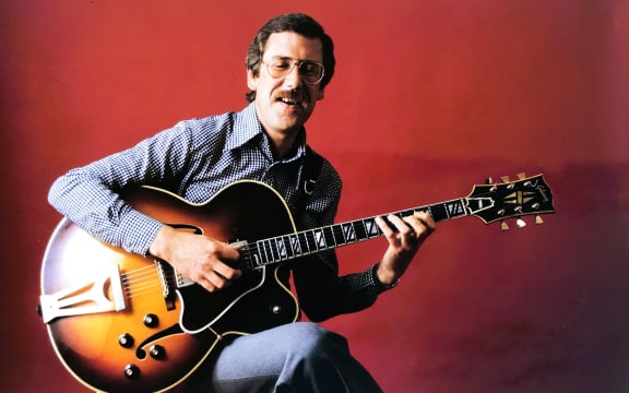 Louis Stewart playing a hollow body guitar. He is sitting on a stool, facing the camera with his eyes closed.