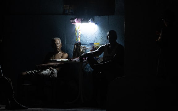 People sit around a table illuminated by a small light source during a nationwide blackout in Havana on July 6, 2026. Cuba on July 6 suffered its third nationwide power outage since the start of the year, the state electricity company said. The impoverished island was already struggling to keep the lights on before US President Donald Trump in January imposed an oil blockade, which has depleted the already dwindling supply of fuel for Cuba's power plants. (Photo by YAMIL LAGE / AFP)