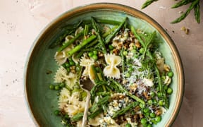 A bowl of farfalle (bow-shaped) pasta with asparagus, peas, blue cheese and walnuts on a pale pink background.