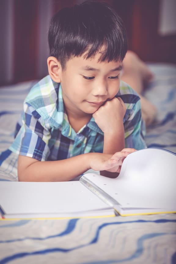 A photo of a young boy reading quietly on his bed. The boy has a look of enjoyment on his face.