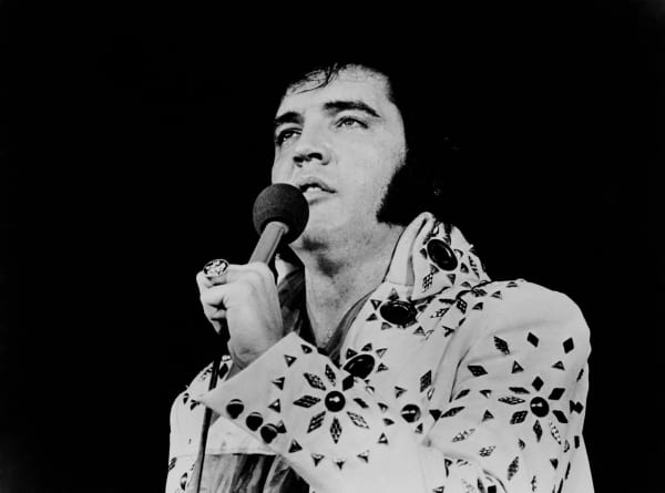 Elvis Presley weaars a white jumpsuit and holds a microphone.E