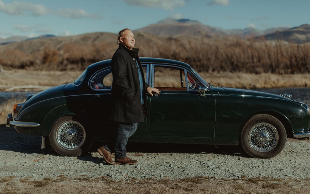 Hamish Bell leans against a vintage Jaguar car in a paddock, wearing a woollen coat, with brown-coloured hills in the background
