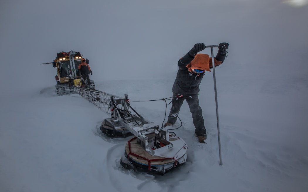 A person in warm gear stands on the ice, wielding a large pole with a T-bar at the top. The person is pushing the pole into the ice. A vehicle with metal contraption attached to it is on the person's right. The scene is dark with grey light, as if the sun is blocked by a thick storm.
