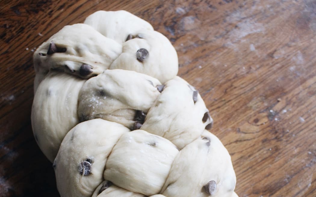 Image of an uncooked loaf of braided chocolate-studded bread on a wooden bread board.