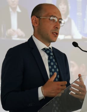 A man wearing a suit, tie and glasses speaking into a microphone with people on a projector screen behind him.