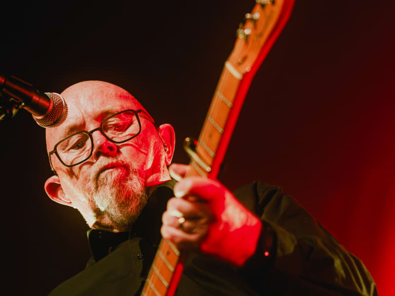 Dave Dobbyn, bathed in red light, looks down at the neck of his guitar.