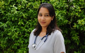 Junior doctor Mourin Das is wearing a stethoscope around her neck and is standing in front of a leafy fence.