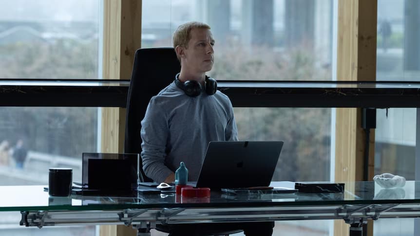 Jeremy Strong as Mark Zuckerberg sitting in a room with a floor-to-ceiling window to his back, sitting with laptop in front and headphones around his neck in The Social Reckoning film.