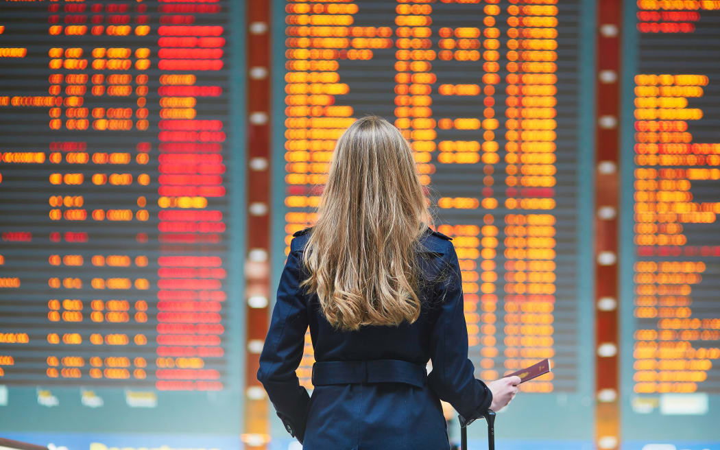 Young woman in international airport looking at the flight information board, holding passport in her hand, checking her flight