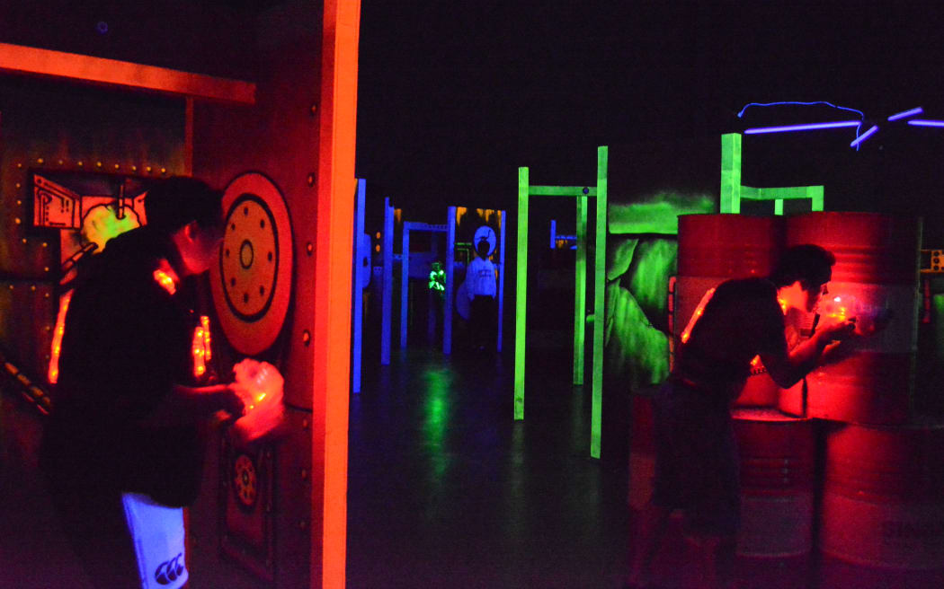 Two people are crouched in action in a laser tag arena. All the lights are out, and walls and weapons are lit up with fluorescent colours.