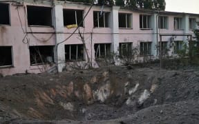 A large hole in the ground in front of a damaged building.