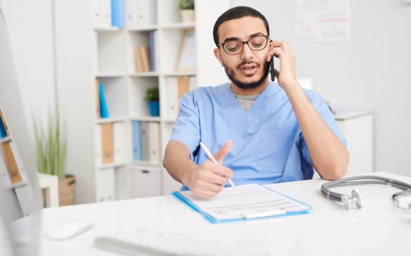 Portrait of young Middle-Eastern doctor wearing glasses sitting at desk in office and speaking by smartphone, filling in patients forms