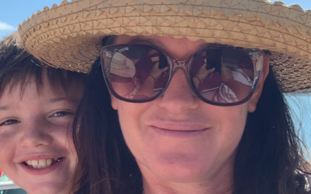 A portrait of Emily in a sun hat and glasses with a boy over her right shoulder, with a backdrop of bright blue sky. She is smiling and wearing a blue polka dot top.
