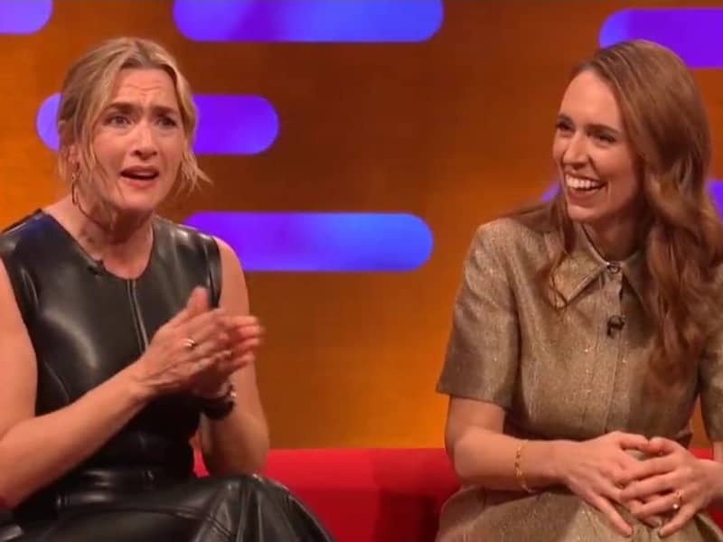 Kate Winslet speaks with clasped hands as Jacinda Ardern smiles and looks on.