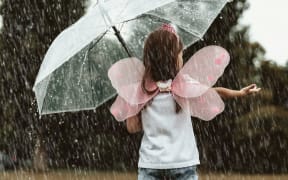Child in fairy wing costume puts hand in rain under umbrella.