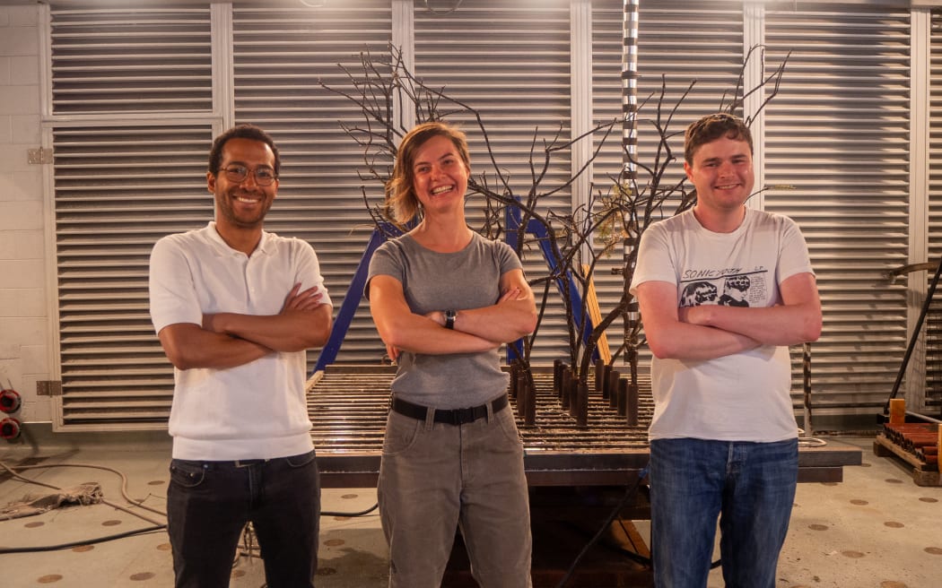 Three people stand with their arms crossed, smiling, in front of a charred bush in a garage-style lab.
