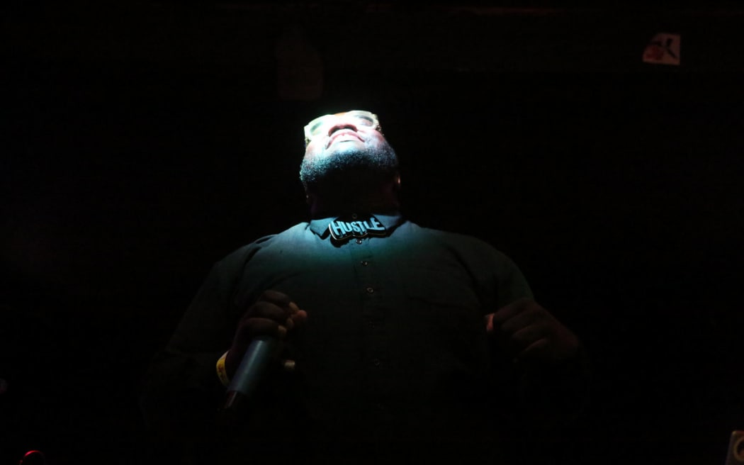 Maga Ran standing in the shadows, his face turned up wards with a spotlight shining on it, wearing a tie that says Hustle.